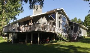 Large house with gray siding wrap around deck and large windows shaded by trees
